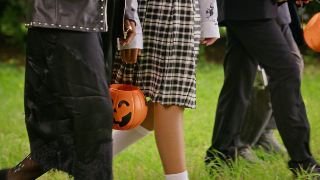 Trick-or-treaters in Halloween costumes walking across a lawn carrying orange pumpkin candy buckets