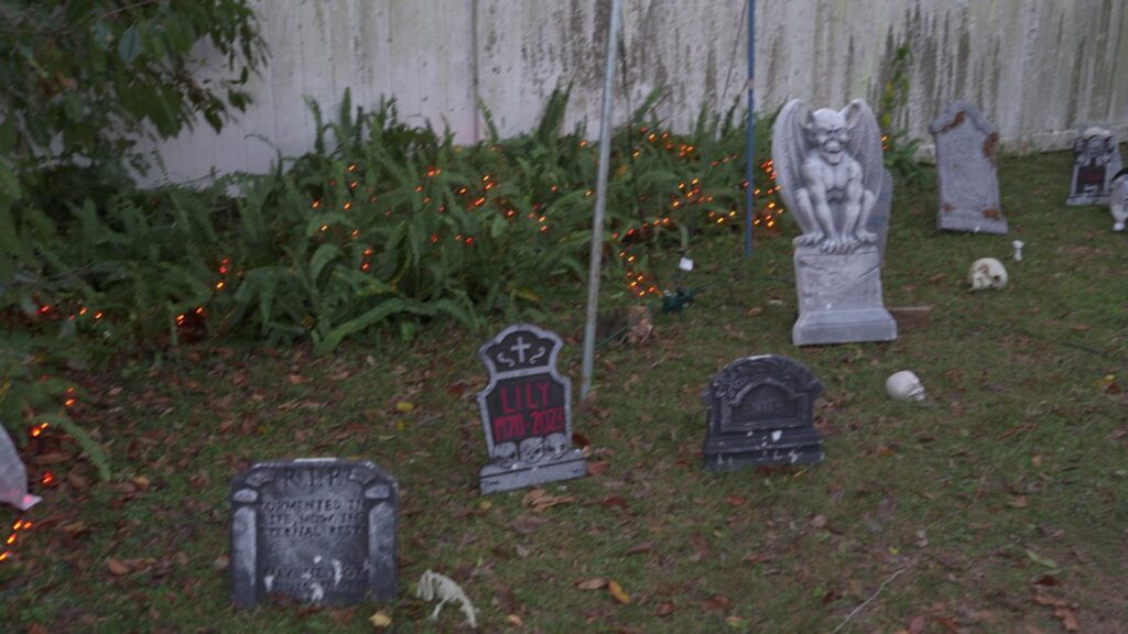 Front-yard Halloween graveyard with foam tombstones, a gargoyle, skulls and orange string lights