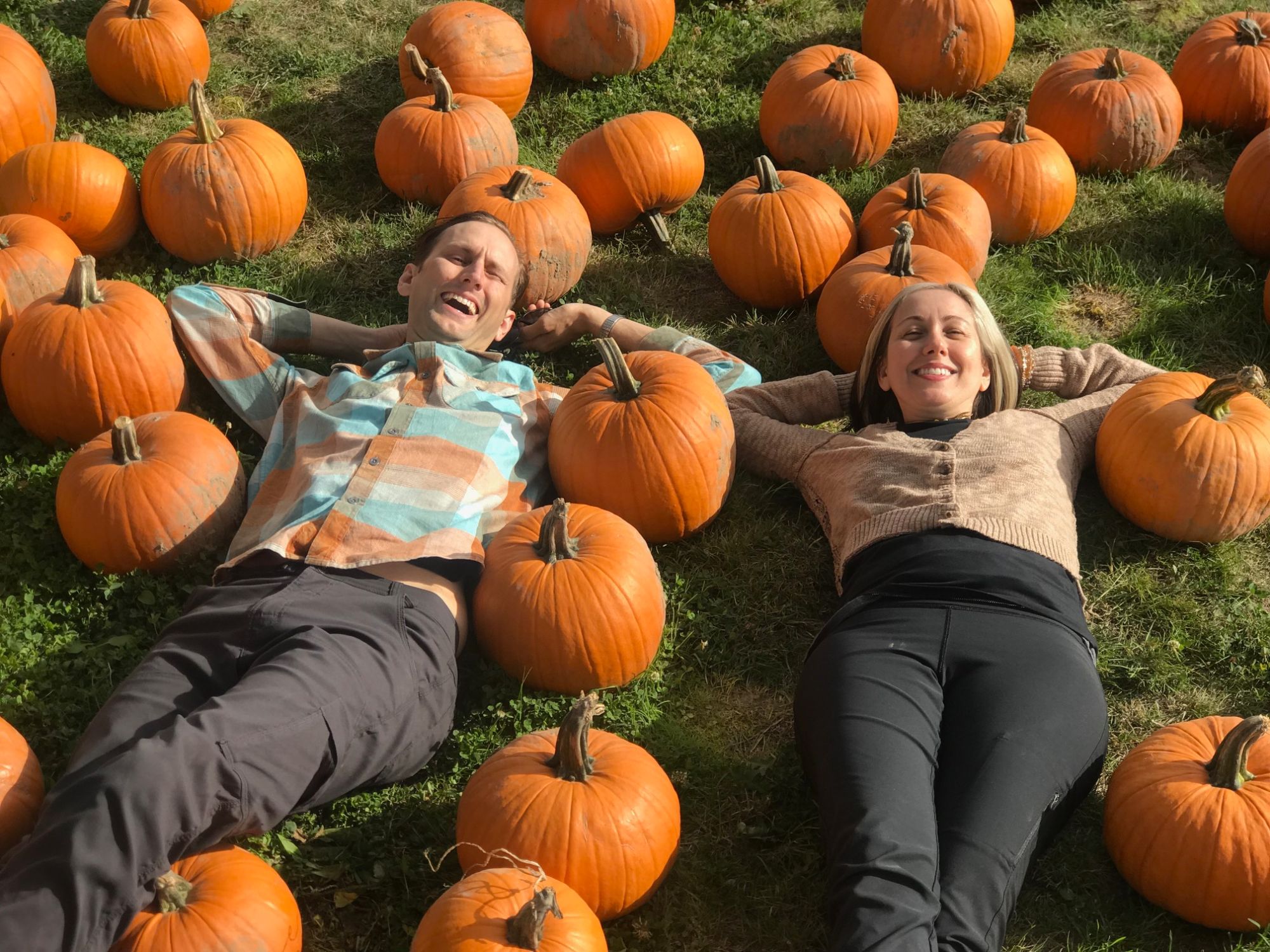 Two people laughing while lying in the grass surrounded by pumpkins at a pumpkin patch