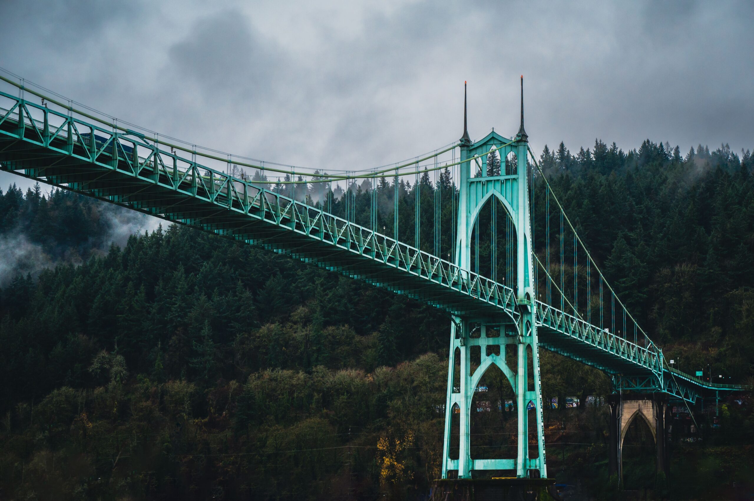Green Gothic arched tower of the St. Johns Bridge in Portland against forested hills and cloudy sky
