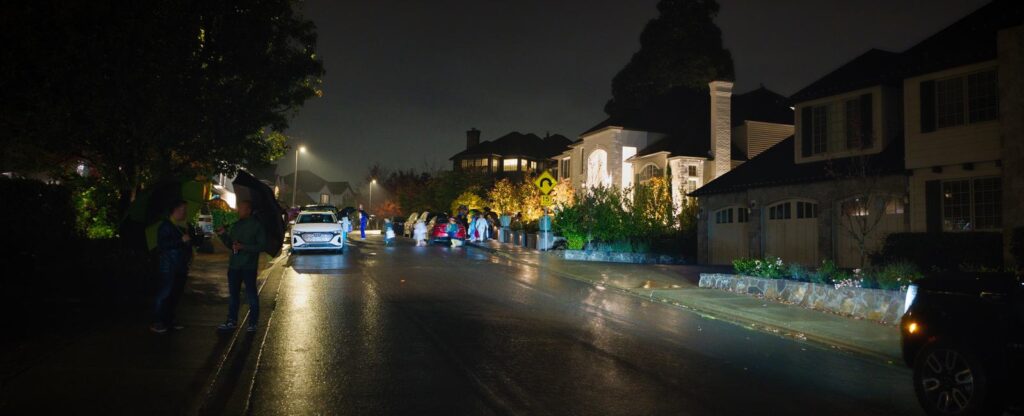Trick-or-treaters with umbrellas walking a wet, lamp-lit residential street on Halloween night